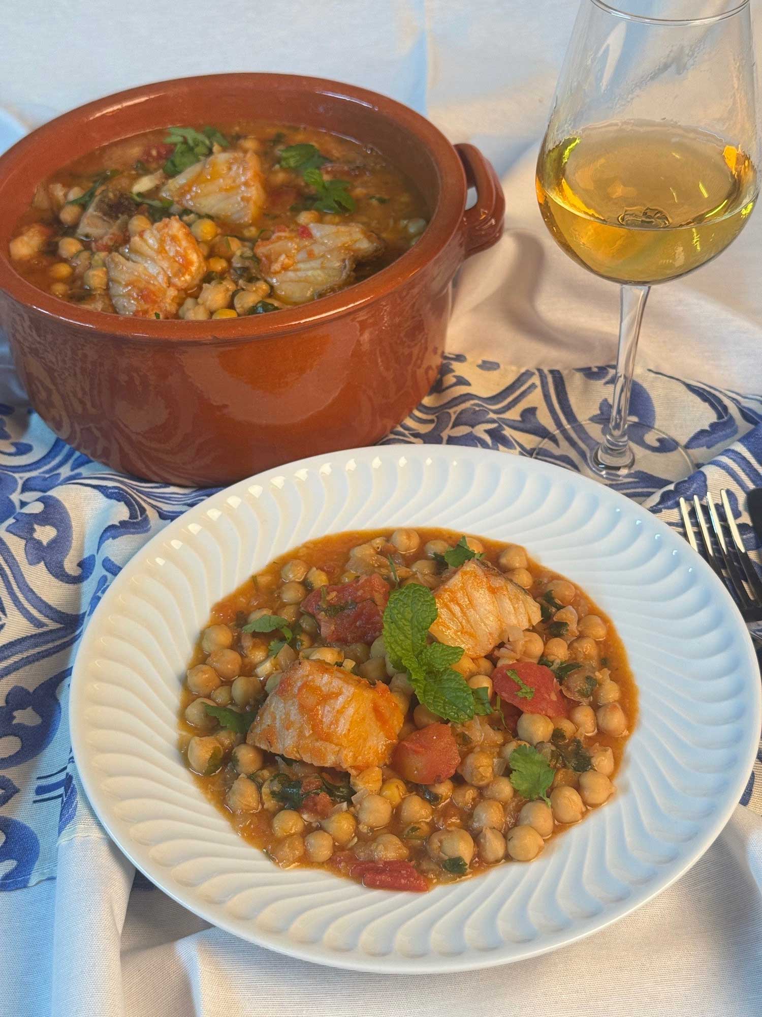 Imagem de Bacalhau guisado com grão num tacho e num prato acompanhado com vinho branco