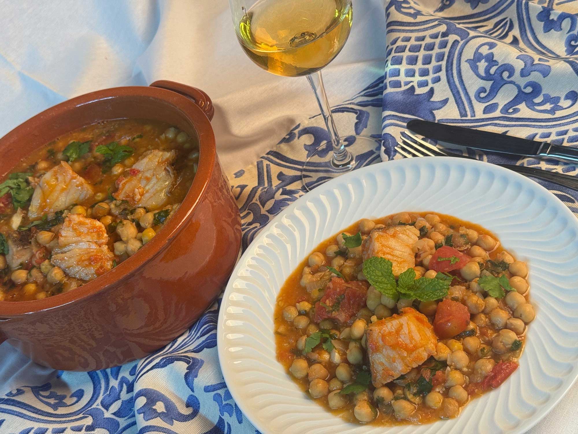 Imagem da receita Bacalhau guisado com grão num tacho de barro e num prato acompanhado com um copo de vinho branco